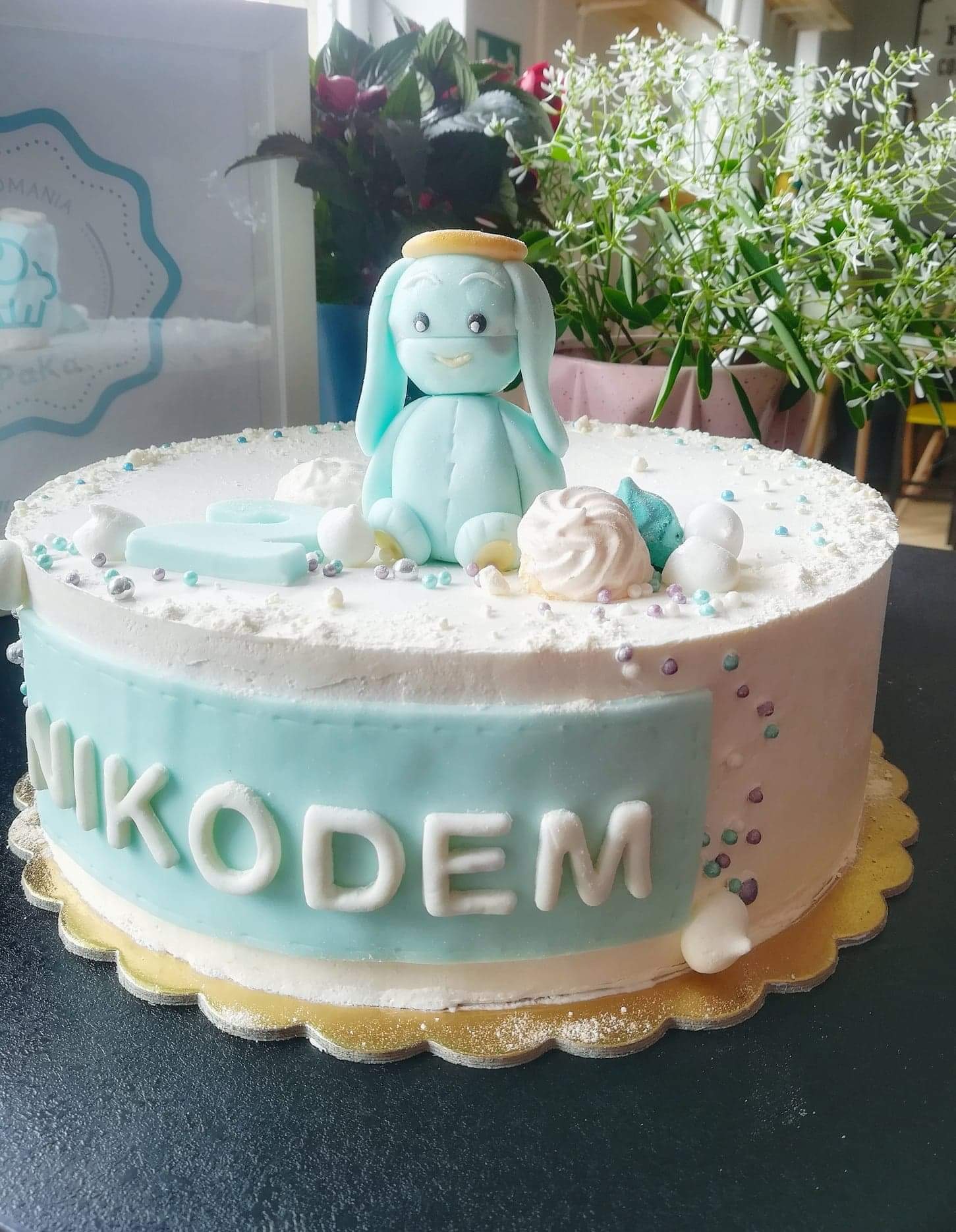 Tort urodzinowy z imieniem 'NIKODEM', udekorowany niebieską figurką królika z aureolą, bezą i kolorowymi posypkami, na złotym podkładzie.