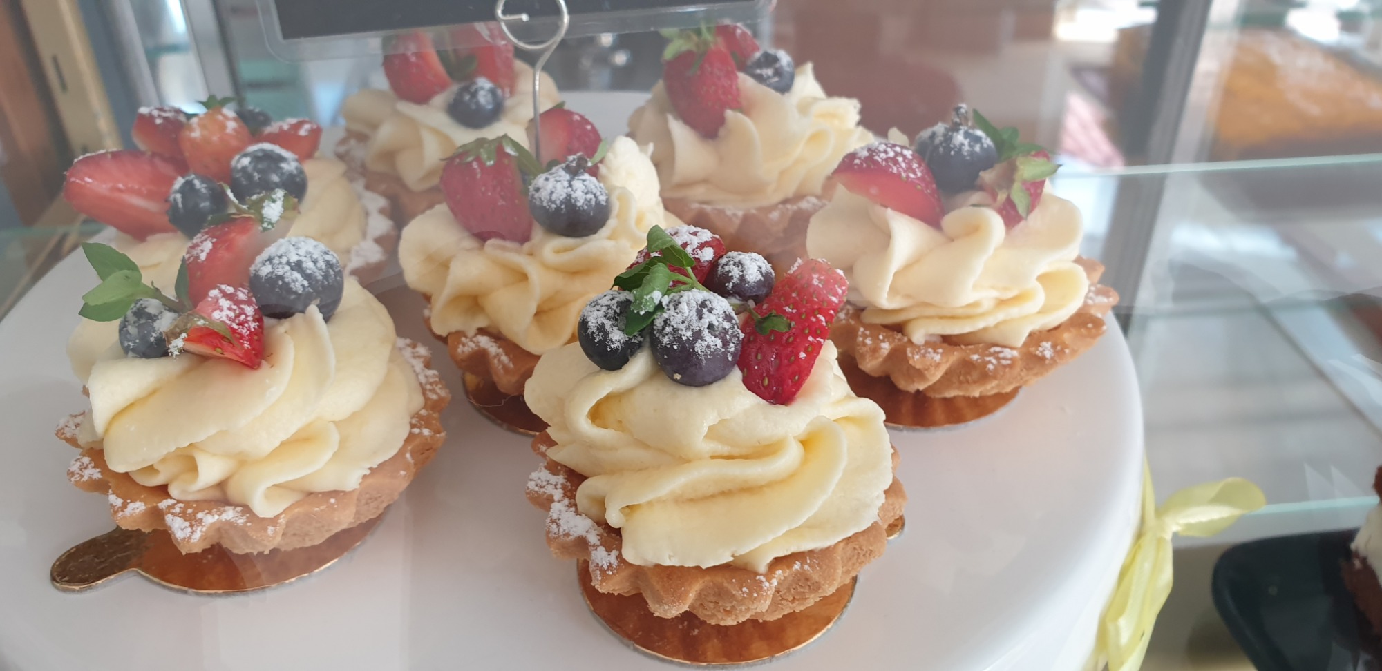 Mini tartaletki z kremem, truskawkami i borówkami, posypane cukrem pudrem, ustawione na białym talerzu w witrynie.