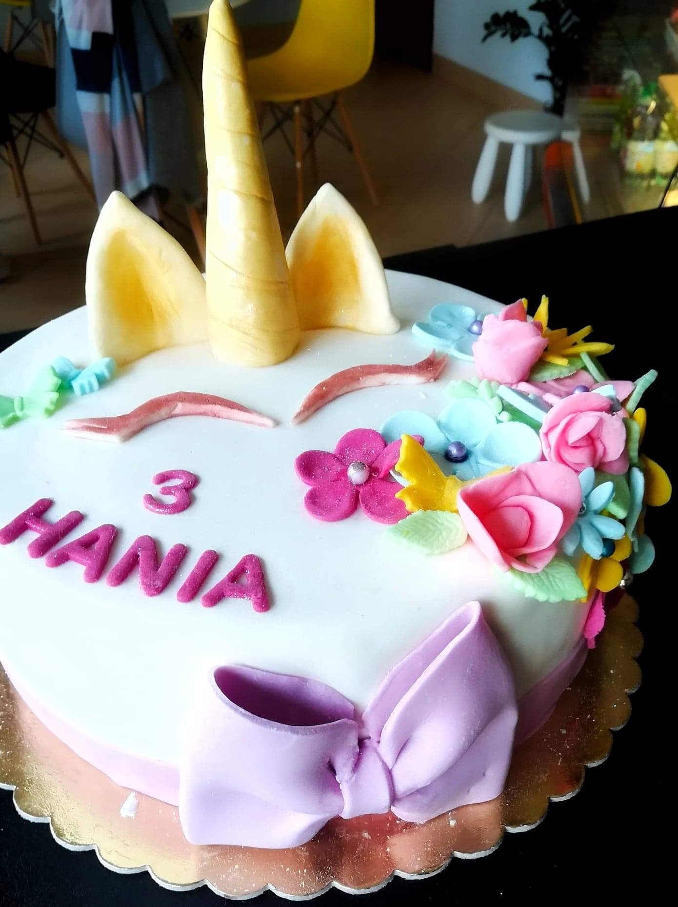 Tort jednorożec z imieniem Hania i cyfrą 3, ozdobiony pastelowymi kwiatami i kokardą, na złotym podkładzie.