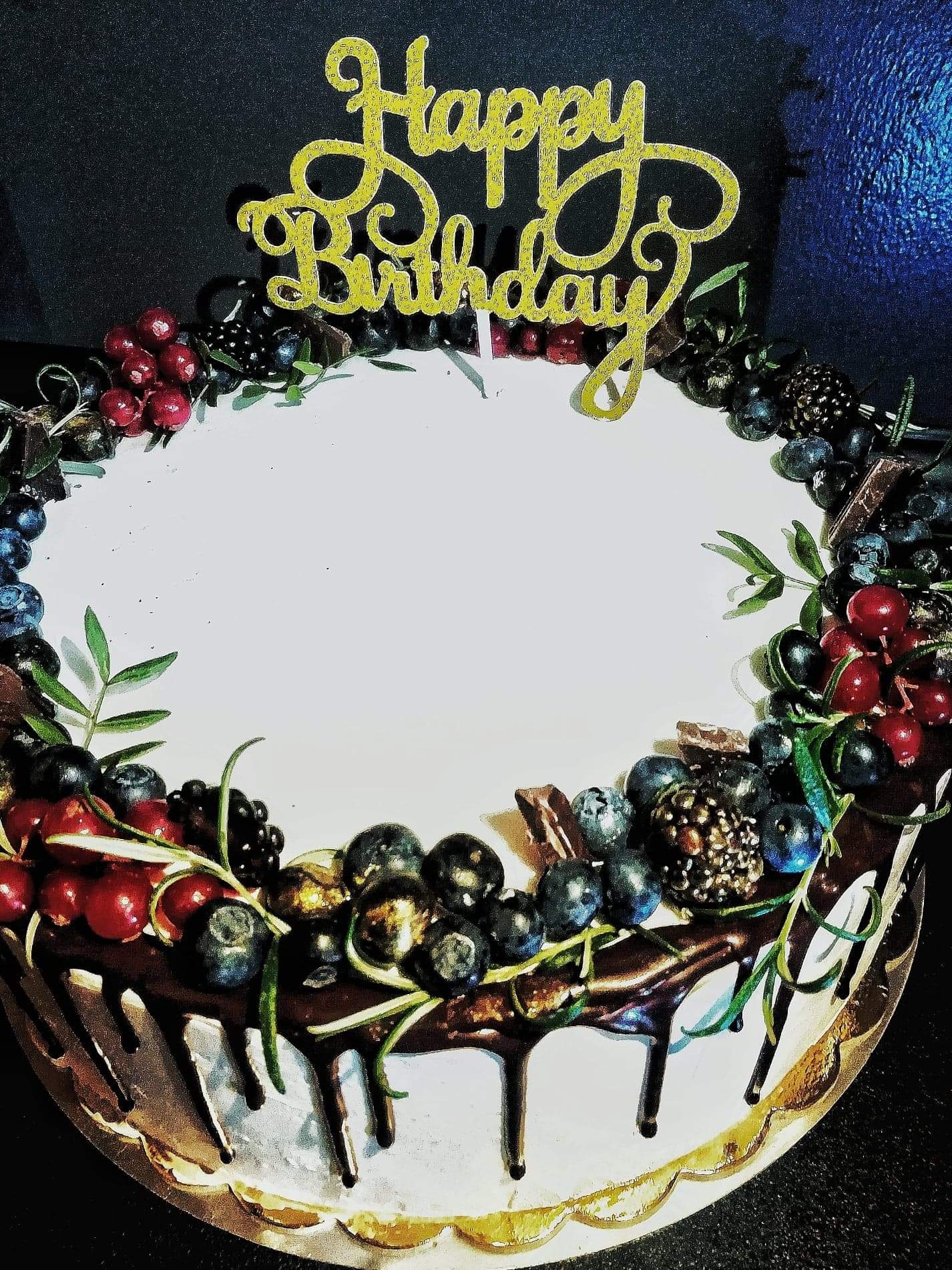 Tort urodzinowy z białym lukrem, obłożony świeżymi owocami leśnymi, kawałkami czekolady i złotym napisem 'Happy Birthday'.
