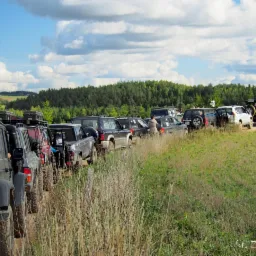 Kolumna samochodów terenowych zaparkowanych na polnej drodze, w tle las i błękitne niebo z obłokami.