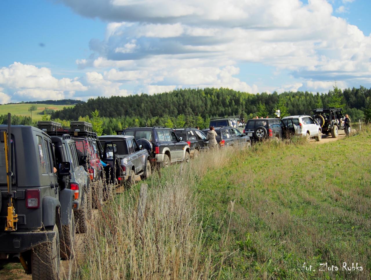Kolumna samochodów terenowych zaparkowanych na polnej drodze, w tle las i błękitne niebo z obłokami.
