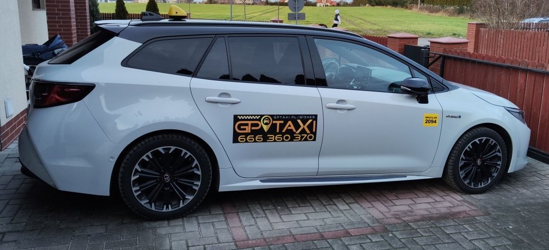 Biała Toyota Corolla kombi z czarnym dachem i oznaczeniami taksówki GP Taxi z numerem telefonu 666 360 370, zaparkowana na kostce brukowej przed brązowym ogrodzeniem.