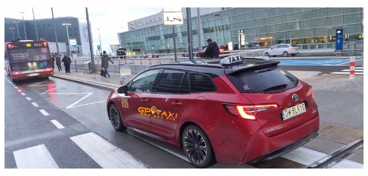 Czerwona taksówka Toyota Corolla kombi z logo GP Taxi i numerem telefonu zaparkowana na postoju taksówek przed lotniskiem Chopina w Warszawie, w tle autobus miejski i budynek terminala.