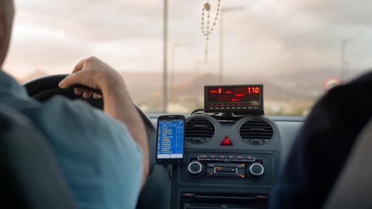 Wnętrze taksówki: widok z tylnego siedzenia na kierowcę trzymającego kierownicę, taksometr z wyświetloną kwotą 7.70, smartfon z listą aplikacji i różaniec wiszący na lusterku.