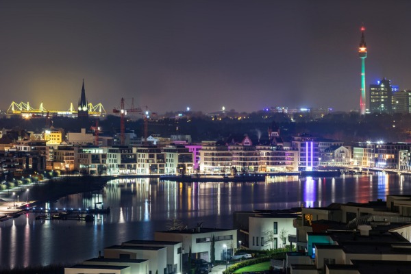 Nocny widok na miasto nad wodą z oświetlonym stadionem i wieżą telewizyjną w tle.