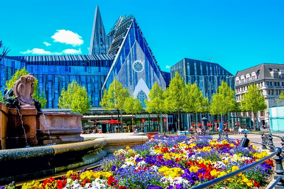 Nowoczesna architektura sakralna z fontanną i klombem kolorowych bratków w pierwszym planie, widok na plac miejski z drzewami i budynkami w Düsseldorfie.