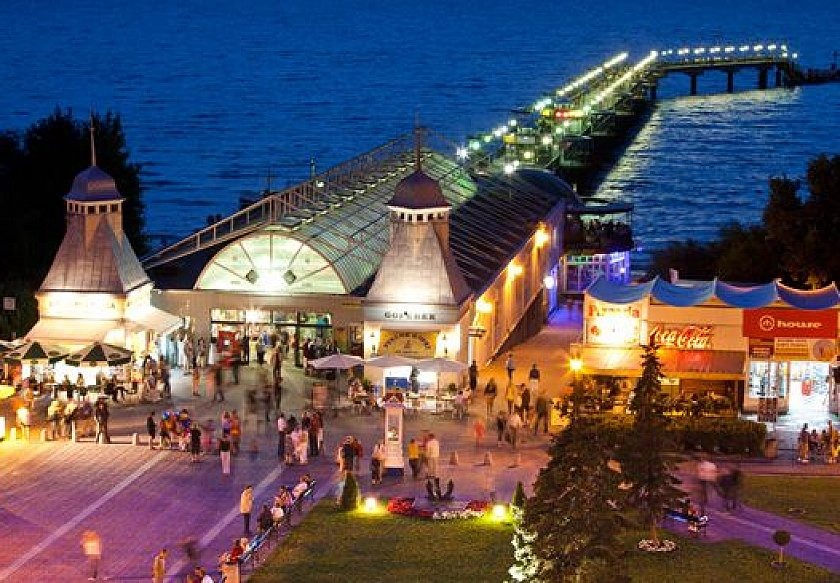 Widok z góry na oświetlone molo w Sopocie z budynkiem wejściowym i promenadą pełną ludzi o zmierzchu.
