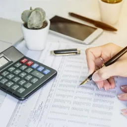 Osoba zaznacza długopisem dane w arkuszu kalkulacyjnym, obok kalkulator, laptop i roślina w doniczce.