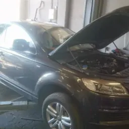 Ciemnoszary SUV Audi Q7 na podnośniku w warsztacie, z otwartą maską ukazującą silnik.