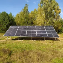 Panel słoneczny ustawiony na metalowej konstrukcji na łące, z drzewami w tle i błękitnym niebem z chmurami.