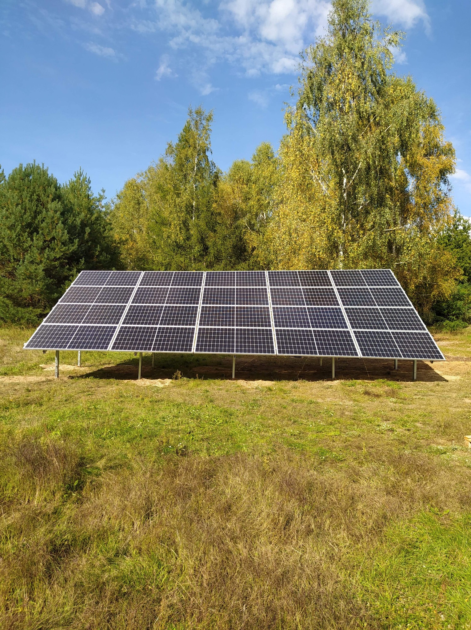 Panel słoneczny ustawiony na metalowej konstrukcji na łące, z drzewami w tle i błękitnym niebem z chmurami.