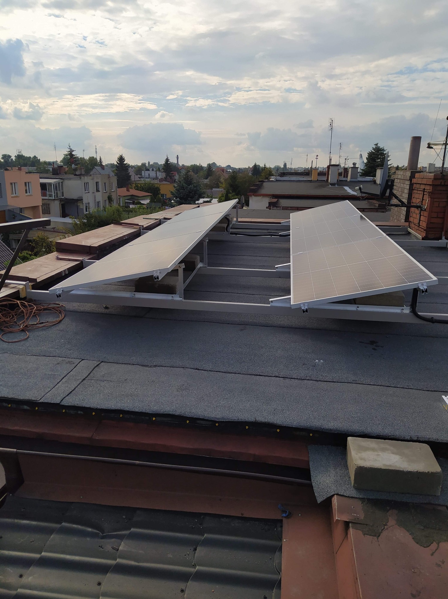 Panele słoneczne na płaskim dachu budynku mieszkalnego, widok z góry, częściowo zamontowane, z widocznym przewodem elektrycznym i elementami mocującymi. W tle zabudowa miejska i pochmurne niebo.