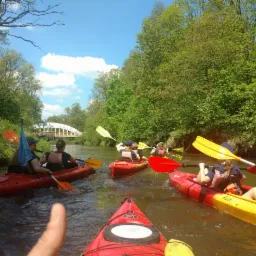 Spływ kajakowy rzeką wśród zieleni, widoczne kajaki z osobami w kamizelkach ratunkowych, wiosła i fragment palca w kadrze.