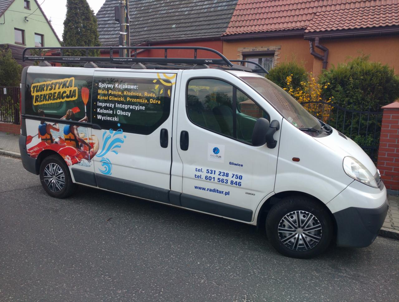 Biały bus z reklamą firmy oferującej spływy kajakowe, imprezy integracyjne, kolonie, obozy i wycieczki, zaparkowany na ulicy.