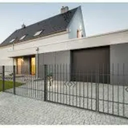 Nowoczesny dom jednorodzinny z szarą elewacją i ciemną bramą garażową, widoczny zza metalowego ogrodzenia z ozdobnymi elementami w kształcie rombów.