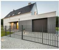 Nowoczesny dom jednorodzinny z szarą elewacją i ciemną bramą garażową, widoczny zza metalowego ogrodzenia z ozdobnymi elementami w kształcie rombów.