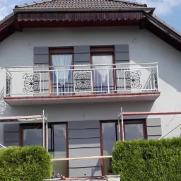 Dwukondygnacyjny dom z szarą elewacją, balkonem z ozdobną balustradą i fragmentem rusztowania przy ścianie bocznej, otoczony zielenią.