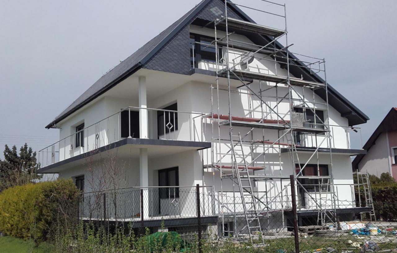 Dwupiętrowy dom z białymi ścianami i szarą dachówką, w trakcie prac wykończeniowych elewacji, z rusztowaniem metalowym przylegającym do ściany, widok z dołu na ukośnej perspektywie.