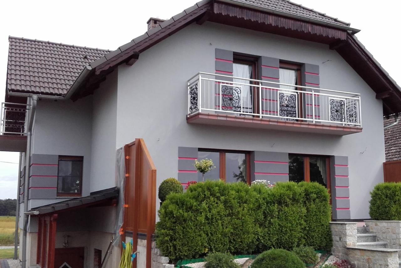 Szary dom jednorodzinny z brązowym dachem, balkonem z ozdobną metalową balustradą, dekoracyjnymi pasami w kolorze fuksji i zadbanym żywopłotem w ogrodzie.