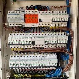 Otwarta skrzynka elektryczna z zamontowanymi bezpiecznikami, wyłącznikami różnicowoprądowymi i przewodami w różnych kolorach, widoczne pomarańczowe zabezpieczenia przeciwprzepięciowe PRO-TEC.