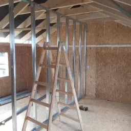 Wnętrze poddasza z drewnianą boazerią i metalowym szkieletem ścian działowych, częściowo ukończone, z drewnianą drabiną i wkrętarką na podłodze.