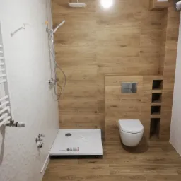 Nowoczesna łazienka z kabiną prysznicową typu walk-in, drewnopodobnymi płytkami na ścianach i podłodze, białą ceramiką i grzejnikiem drabinkowym, widoczna tuba silikonu sanitarnego na brodziku.