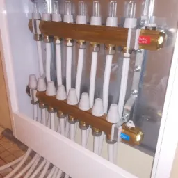 Rozdzielacz ogrzewania podłogowego w białej szafce, widoczne zawory regulacyjne, rury doprowadzające i odprowadzające wodę oraz przepływomierze