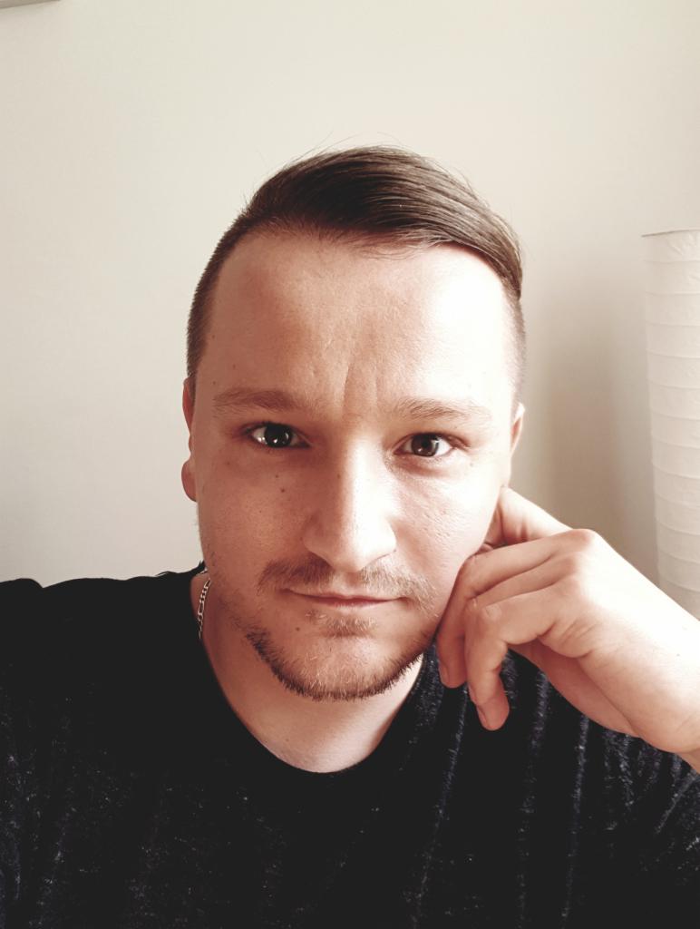 Portret mężczyzny z krótką fryzurą i lekkim zarostem, opierającego brodę na dłoni, z zamyślonym wyrazem twarzy i delikatnym uśmiechem.