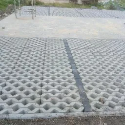 Ułożona szara ażurowa kostka brukowa na podjeździe z widocznymi elementami konstrukcji w tle.