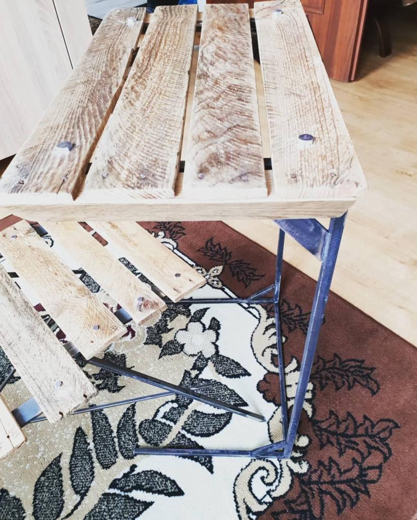 Stolik kawowy z blatem z desek o widocznej strukturze drewna, osadzony na metalowej ramie, na tle wzorzystego dywanu.