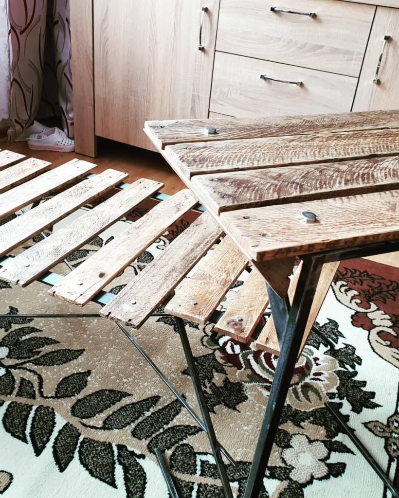 Rozłożony blat drewnianego stolika kawowego z metalowym stelażem na wzorzystym dywanie, widoczne elementy mocujące i niedokończone deski blatu, w tle fragment szafki i osoba w białych butach.