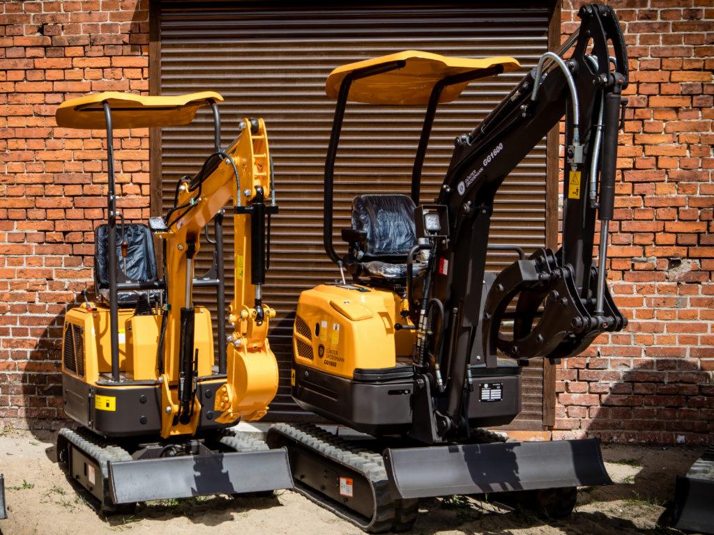 Dwie nowe, żółto-czarne minikoparki stoją obok siebie na tle ceglanej ściany i zamkniętych metalowych drzwi garażowych. Jedna z nich ma zamontowane chwytaki zamiast łyżki.