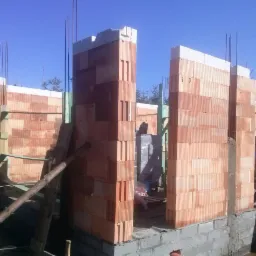 Wznoszenie ścian domu z czerwonej cegły ceramicznej na fundamencie z betonowych bloczków, widoczne zbrojenia pionowe i drewniane podpory, w tle drzewa i błękitne niebo.