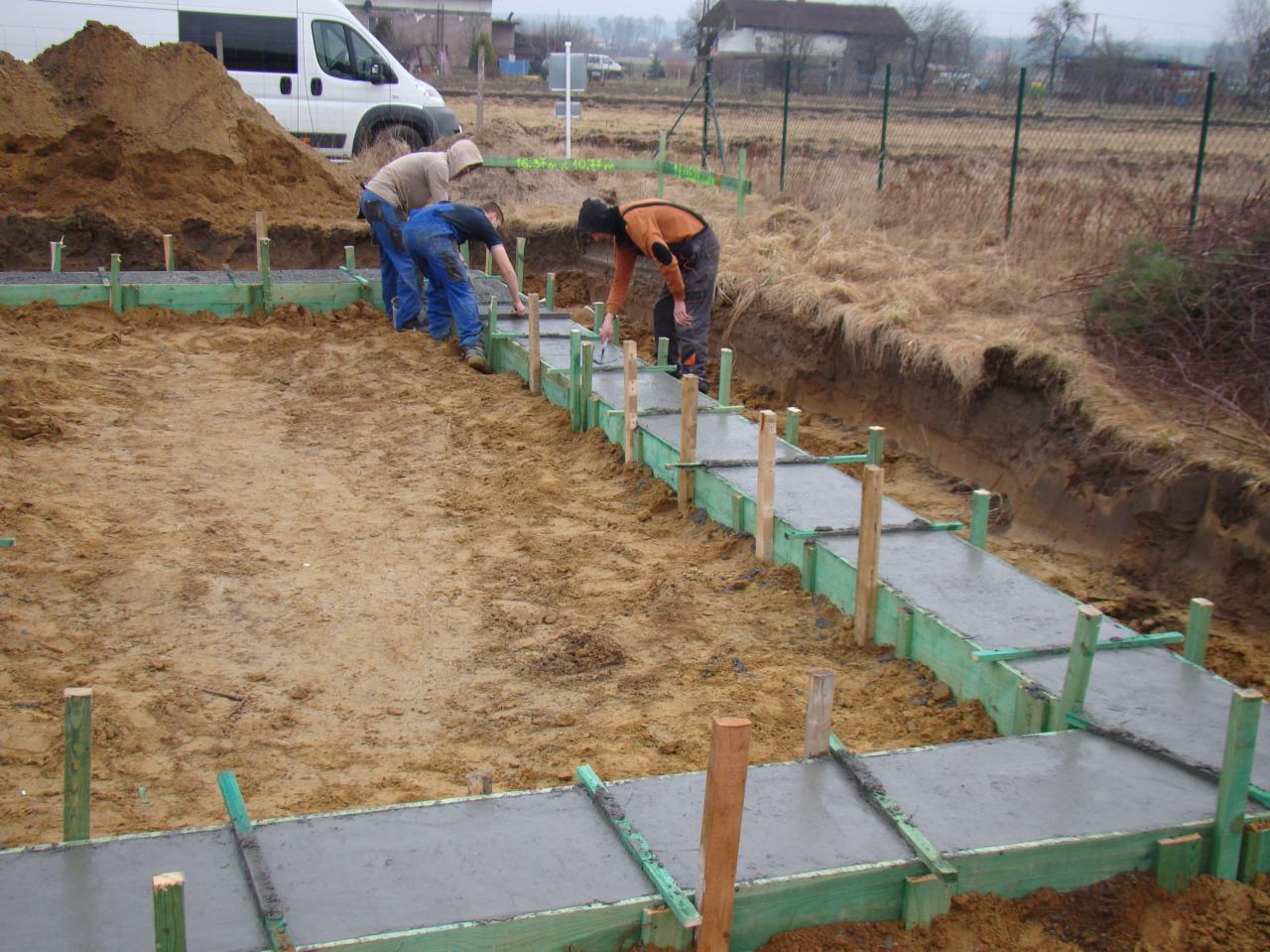 Trzech pracowników wyrównuje świeżo wylaną wylewkę fundamentową w szalunkach z zielonych desek, na tle wykopu, sterty ziemi i zabudowań.