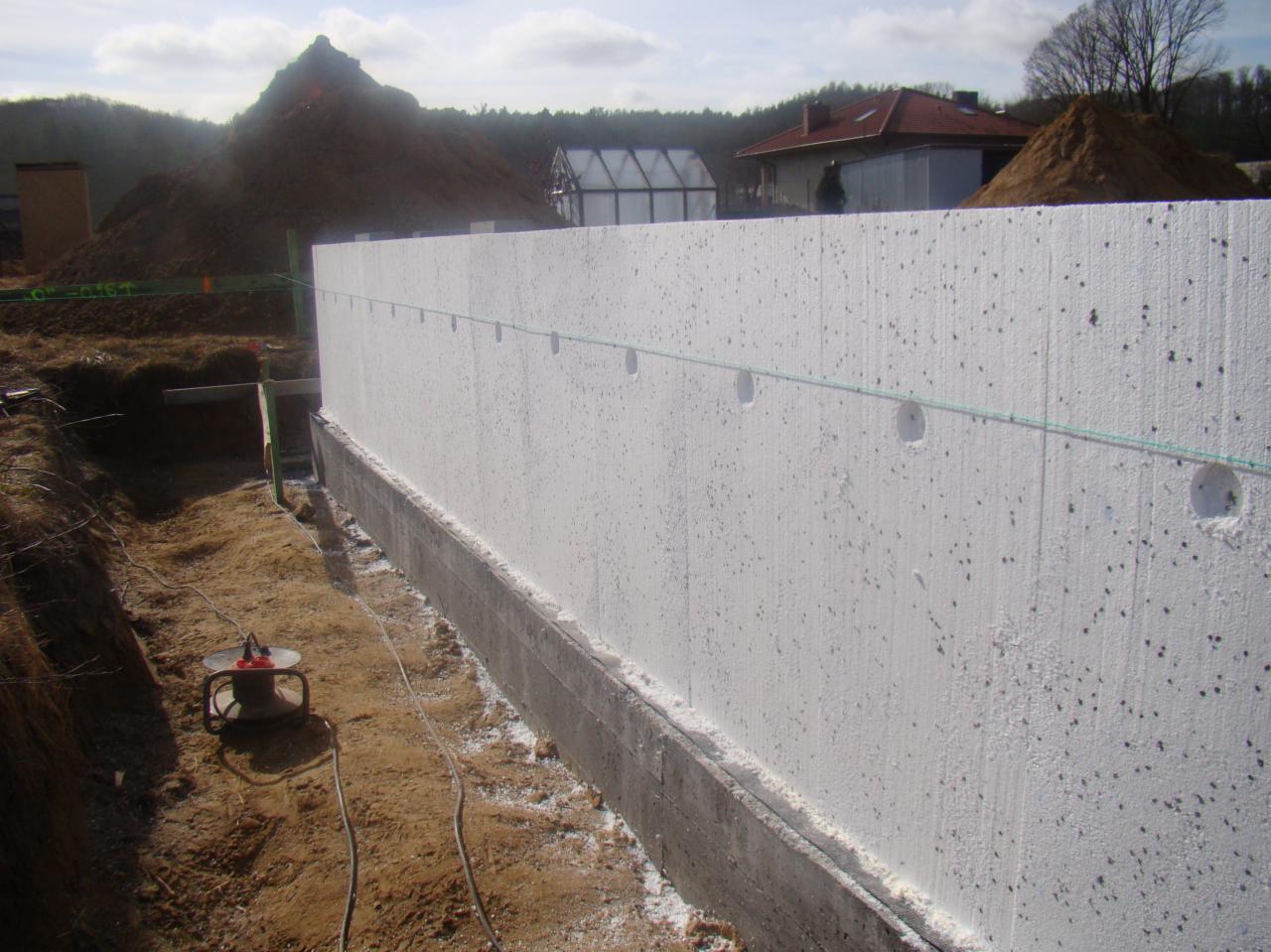 Fundament budynku ocieplony styropianem, widoczne otwory na kotwy, w tle ziemia i budynki w budowie.