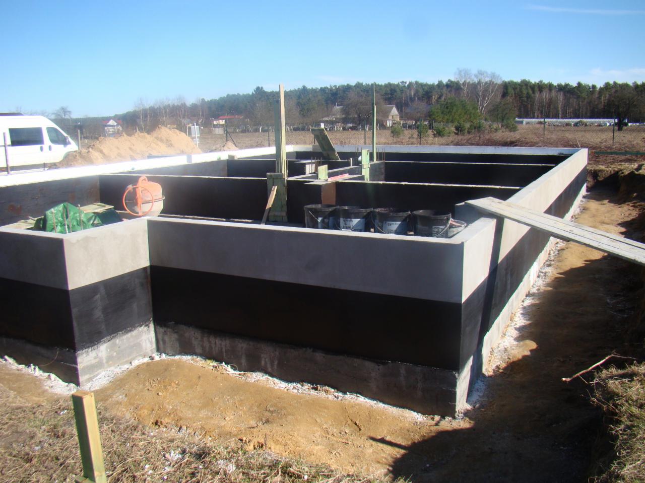 Fundamenty domu z hydroizolacją bitumiczną, widoczne szalunki, mieszalnik betonu i wiaderka z materiałami budowlanymi, w tle las i błękitne niebo.