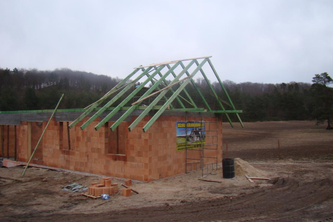 Wznoszenie budynku z czerwonej cegły z widoczną konstrukcją drewnianą dachu na tle zalesionego wzgórza i pochmurnego nieba.