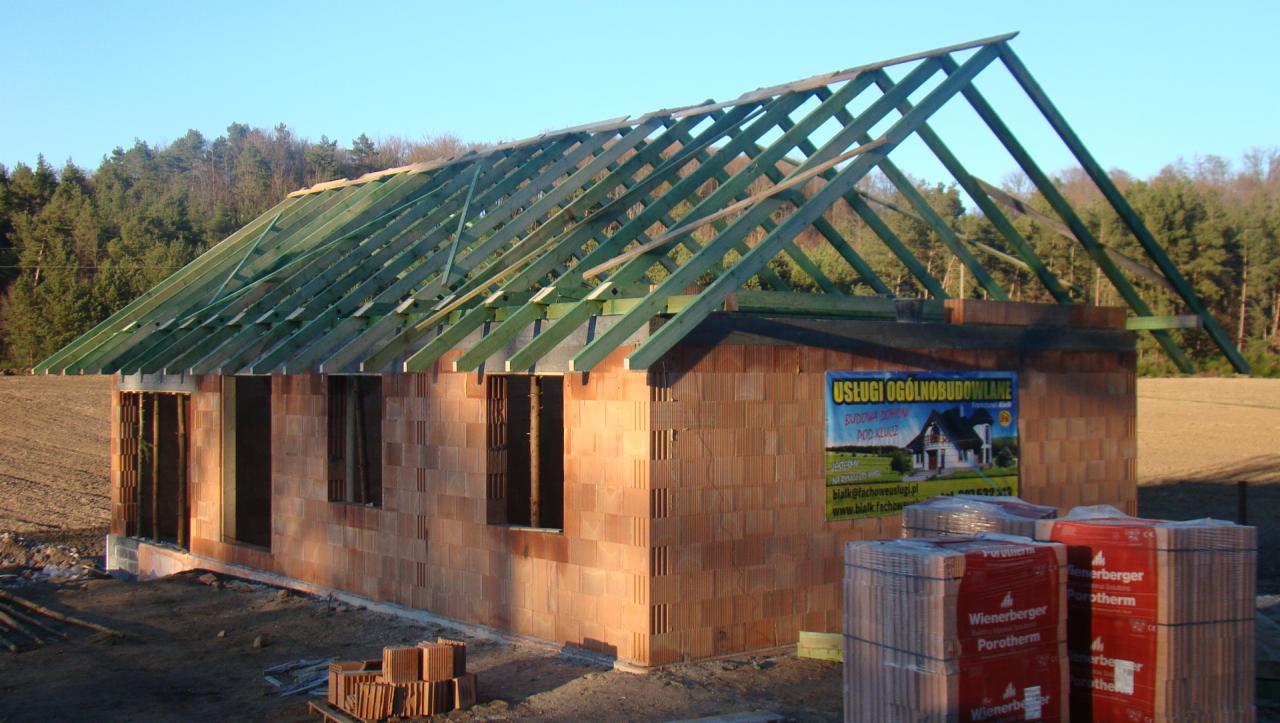 Wznoszony budynek z pustaków ceramicznych Porotherm, z widoczną konstrukcją więźby dachowej z impregnowanego drewna; na ścianie budynku baner reklamowy usług ogólnobudowlanych, a przed budynkiem...