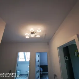 Nowoczesna lampa sufitowa LED z okrągłymi elementami w jasnym korytarzu z dwoma drzwiami i lukiem rewizyjnym na suficie.