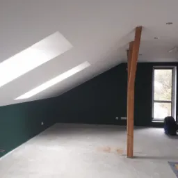Poddasze po częściowym remoncie: ściany pomalowane na ciemnozielono, biały sufit ze skośnymi oknami, drewniany słup konstrukcyjny, betonowa podłoga, drabina oparta o ścianę przy oknie.