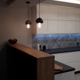 Nowoczesna kuchnia z białymi szafkami, czarnym blatem i dekoracyjnymi płytkami nad blatem, oświetlona wiszącymi lampami z żarówkami Edison i listwą LED pod szafkami.