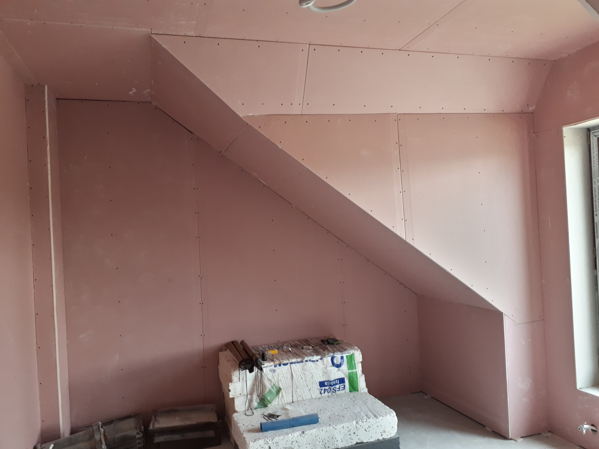 Poddasze z zamontowanymi płytami kartonowo-gipsowymi, widoczne narzędzia i materiały budowlane, okno w rogu pomieszczenia.