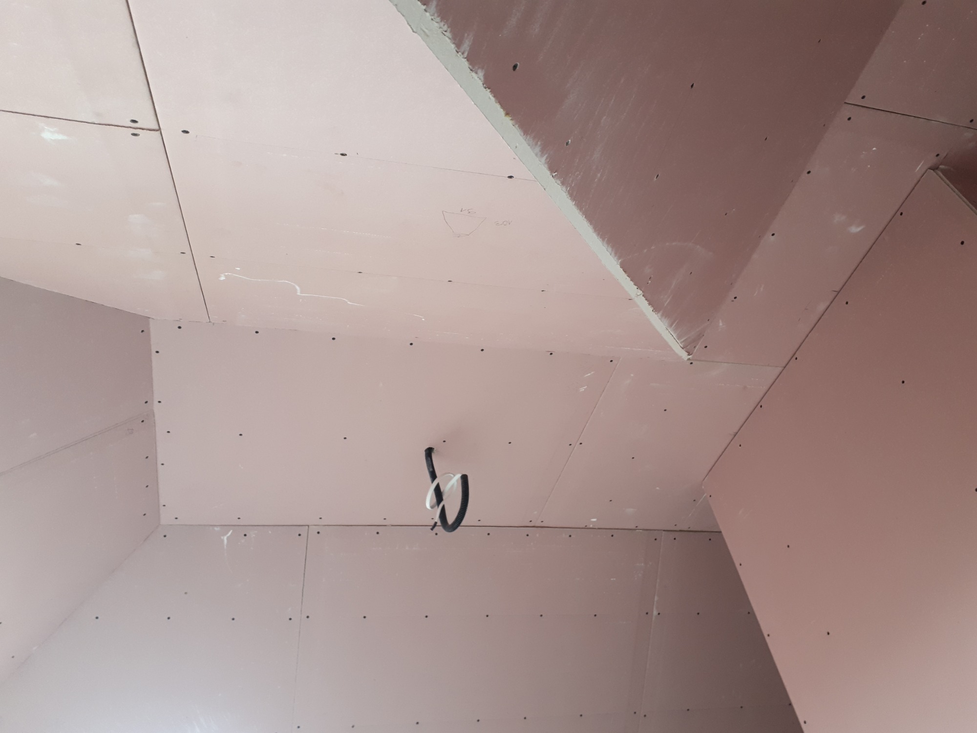 Surowe wnętrze poddasza wyłożone płytami kartonowo-gipsowymi, widoczne łączenia płyt i przewody elektryczne zwisające z sufitu, przygotowane do dalszych prac wykończeniowych.