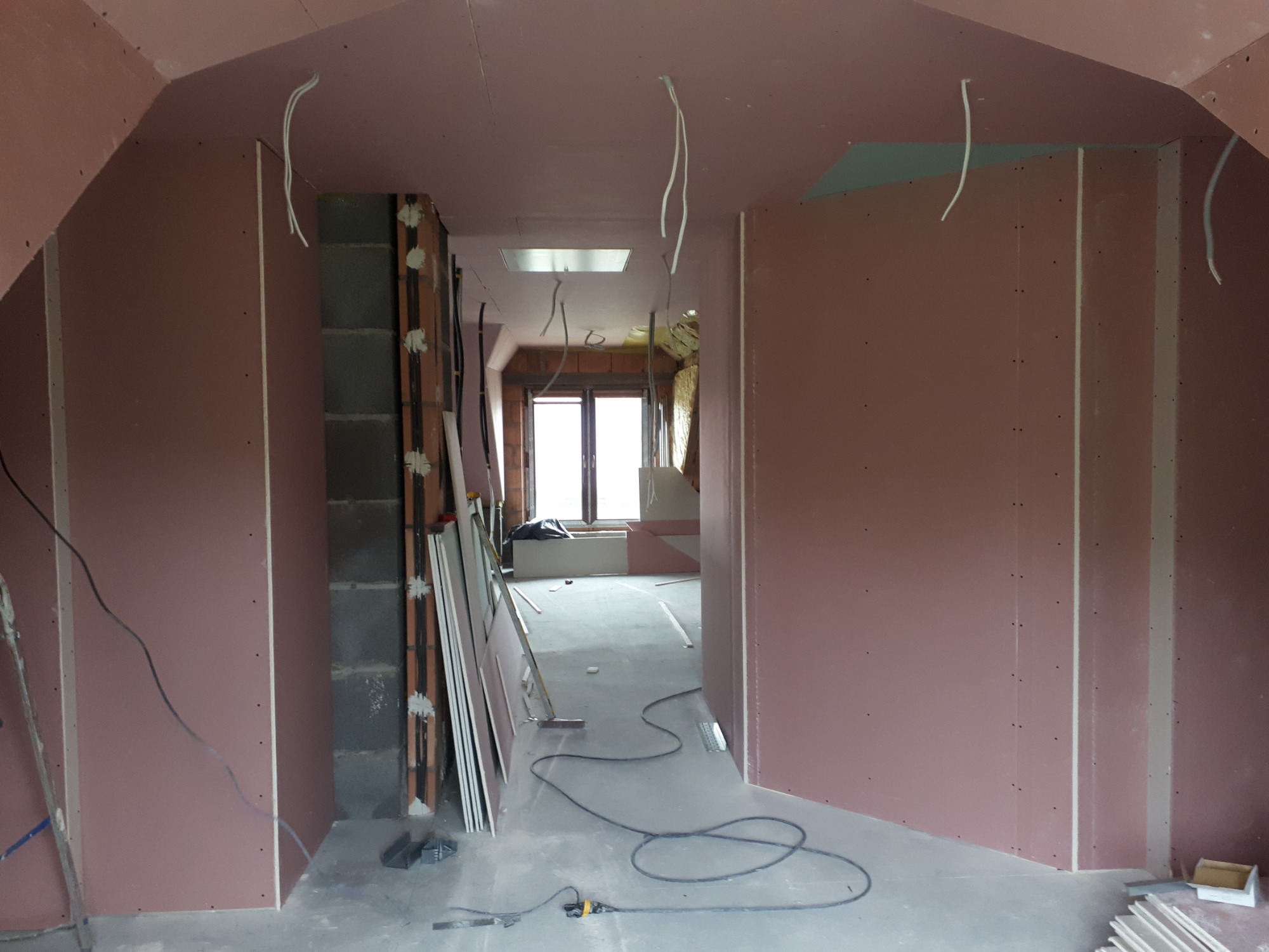 Montaż płyt kartonowo-gipsowych na poddaszu, widoczne profile, kable i otwór okienny w oddali.