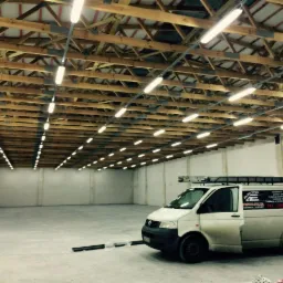 Równomiernie rozmieszczone lampy jarzeniowe pod drewnianą konstrukcją dachu w hali. Biały bus elektryczny z logo firmy i drabina na dachu stoi w rogu hali.