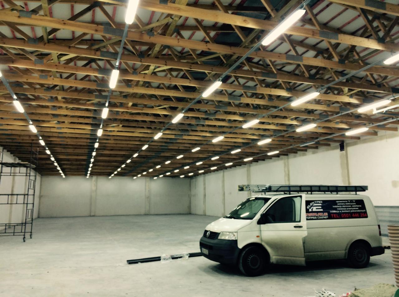 Równomiernie rozmieszczone lampy jarzeniowe pod drewnianą konstrukcją dachu w hali. Biały bus elektryczny z logo firmy i drabina na dachu stoi w rogu hali.