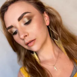 Portret kobiety z brązowo-złotym makijażem oczu, błyszczącymi ustami i złotymi kolczykami w kształcie rombów z frędzlami, okulary przeciwsłoneczne na głowie.