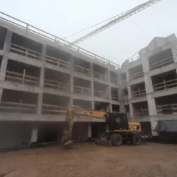 Surowy szkielet betonowy budynku wielopiętrowego w trakcie budowy, widoczny żółty koparko-ładowarka CAT i żuraw w tle, pochmurny dzień.