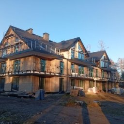 RKON Projekty i nadzór w budownictwie ROGALA KAMIL - Wielorodzinny budynek mieszkalny w zaawansowanej fazie budowy, z jasną elewacją, ciemnym dachem, balkonami z balustradami i rusztowaniami na poddaszu, na nieutwardzonym terenie.